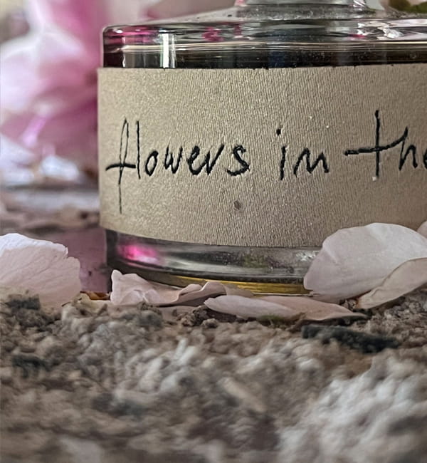 Ilustracja dla zapachu Flowers in the ashes. Zbliżenie butelki perfum z żakardową etykietą stojącej w popiele.